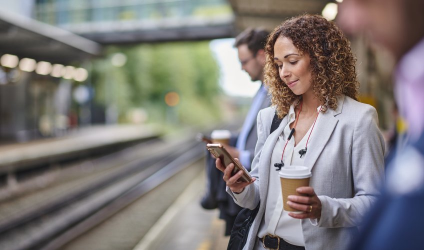 Businesswomen waiting for train on her morning commute looking at her phone holding a cup of coffee.
