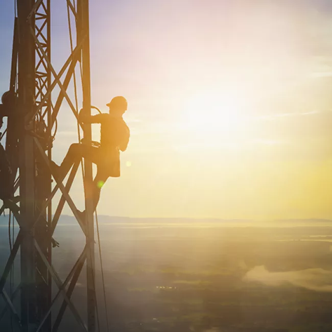 Silhouette of two people working on an electrical tower with the sun and aerial view of hills.